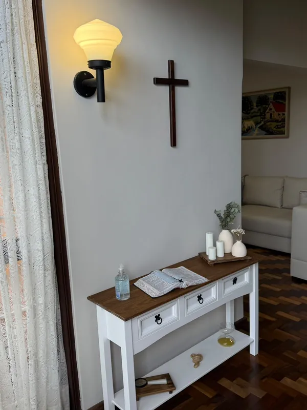 Espaço ecumênico com crucifixo de madeira, aparador branco, velas e uma pequena planta.