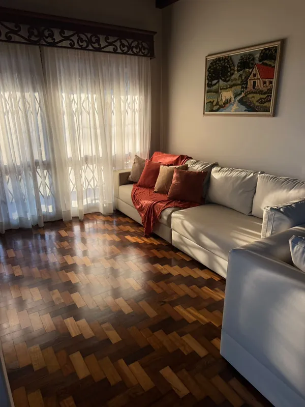 Sala de estar com sofás brancos, almofadas em tons terrosos, cortinas claras e piso de taco.
